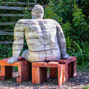 Eine Steinskulptur von einem Mann, der auf einer Holzbank sitzt, mit dem Rücken zum Betrachter und auf ein Gebüsch starrt.