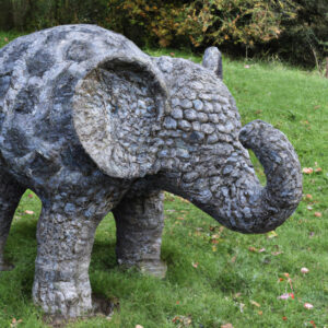 Eine graue Steinskulptur eines Elefanten mit schuppenartiger Oberfläche steht auf einer grünen Wiese, umgeben von Bäumen.