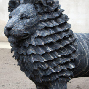 Eine monumentale, dunkelgraue Steinskulptur eines Löwen mit mehrfach geschichteter Mähne, die wie Gesteinsformationen wirkt.