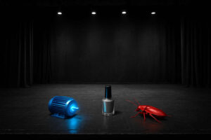 Eine schwarze Theaterbühne mit Spotlights zeigt drei rätselhafte Objekte: einen leuchtend blauen Motor, einen silbernen Nagellack und eine knallrote Kakerlake – jedes einzeln in Szene gesetzt.
