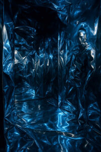 Dunkler Raum mit blau-metallisch reflektierenden Wänden, in denen sich eine schemenhafte Figur spiegelt.