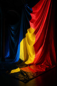 Riesige Stoffbahnen in Blau, Gelb und Rot hängen dramatisch in einem dunklen Raum. Das Licht fällt so, dass die Falten der Stoffe plastisch hervortreten. Das Bild wirkt feierlich, fast sakral, und lässt die Farben als Protagonisten erscheinen.