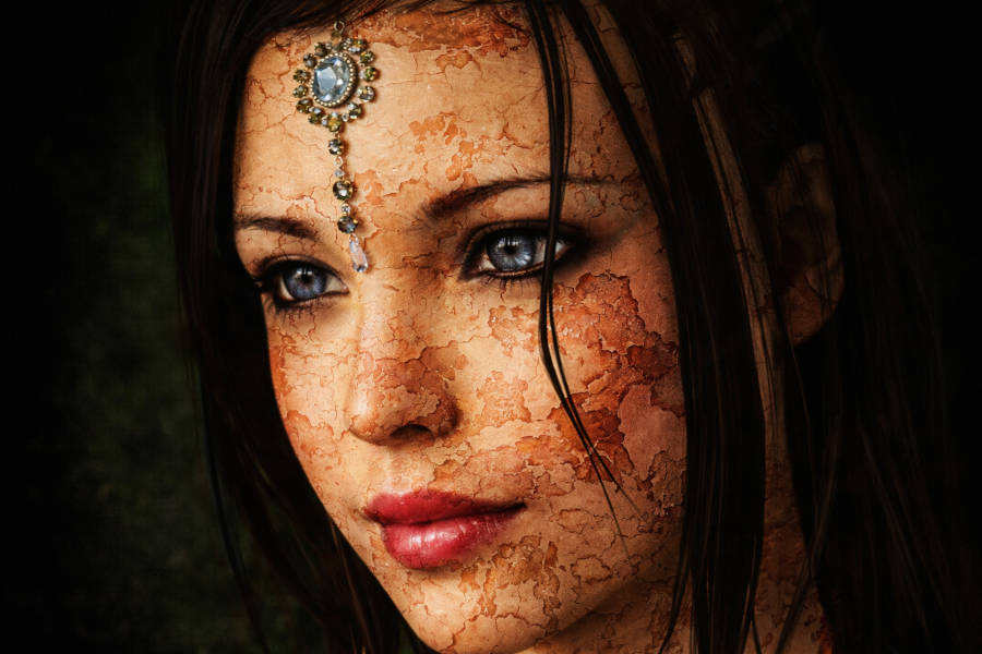 „Detailreiches Porträt einer weiblichen, göttlich anmutenden Figur mit blau-grauen Augen, dunklem Haar und einem Schmuckstein auf der Stirn; ihre Haut wirkt rissig und erdig wie getrockneter Lehm, vor dunklem Hintergrund.“