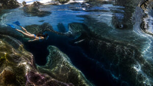 Taucherin schwebt über einer gewaltigen Unterwasserspalte, deren Tiefe im Dunkel verschwindet. Das klare Wasser erlaubt den Blick auf bizarre Felsformationen und geheimnisvolle Schatten. Lichtreflexe tanzen über die Oberfläche und erzeugen ein Gefühl von Schwerelosigkeit. Die Szene verbindet Schönheit mit einer leisen Spannung des Unbekannten.