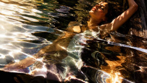 Im warmen Abendlicht treibt eine Frau entspannt im Wasser neben einem hölzernen Steg. Lichtreflexe brechen sich in den Wellen und erzeugen goldene, abstrakte Muster auf der Oberfläche. Die Szene strahlt Ruhe und körperliche Gelöstheit aus. Wasser, Licht und Spiegelungen bilden gemeinsam den eigentlichen Mittelpunkt des Bildes.