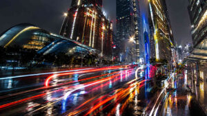 Moderne Großstadt bei Nacht mit spiegelnden Straßen und farbigen Lichtspuren vorbeifahrender Fahrzeuge. Hochhäuser ragen in den dunklen Himmel und reflektieren Blau-, Rot- und Goldtöne. Die nasse Fahrbahn verstärkt die Leuchtkraft der Szene durch zahllose Spiegelungen. Das Bild vermittelt Dynamik, Geschwindigkeit und urbane Energie.