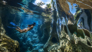 Taucherin gleitet durch ein Labyrinth aus Wasser, Felsen und spiegelnden Formationen. Die Oberflächen verzerren die Umgebung zu flüssigen Reflexionen und erzeugen einen surrealen Eindruck. Sonnenlicht dringt durch das Wasser und beleuchtet die Unterwasserlandschaft in kräftigen Blautönen. Das Bild vermittelt Freiheit, Entdeckung und räumliche Tiefe.