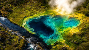 Luftaufnahme einer intensiv türkisfarbenen heißen Quelle mit tiefem dunklem Zentrum. Umgeben von moosbewachsenen Felsen, dampfenden geothermalen Feldern und einem Bachlauf. Die leuchtenden Mineralablagerungen erzeugen starke Farbkontraste zwischen Grün, Blau und Gelb. Die Szene wirkt wie ein verborgenes Naturwunder aus einer fremden Welt.