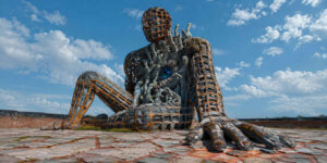 Eine monumentale sitzende Menschengestalt erhebt sich auf einem gepflasterten Platz unter blauem Himmel. Ihr steinerner Körper wird von einem orangefarbenen Gitter zusammengehalten. Aus dem geöffneten Rumpf drängen zahlreiche Hände und Figuren hervor, zwischen denen ein großes blaues Auge sichtbar wird. Zwei winzige Menschen stehen vor ihrem angewinkelten Bein.