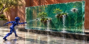 Eine blaue, kristallartig glänzende Figur wirft einen kleinen dunklen Stein gegen eine große transparente Wasserwand. Innerhalb dieser Wand schweben mehrere kleine Inseln mit Bäumen, Moos und Wasserfällen, die sich wie Miniaturlandschaften im Wasser halten. Von der Auftreffstelle breiten sich deutliche Wellenkreise aus. Rechts oben wächst aus einer weiteren kleinen Insel bereits ein frischer grüner Keimling.