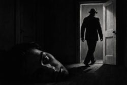 Eine riesige Männerfigur liegt mit dem Kopf auf dem Boden, ausdruckslos. Im Hintergrund verlässt ein anderer Mann mit Hut das Zimmer durch eine offene Tür ins Licht. Die Szene ist in Schwarz-Weiß gehalten.