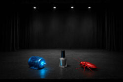 Eine schwarze Theaterbühne mit Spotlights zeigt drei rätselhafte Objekte: einen leuchtend blauen Motor, einen silbernen Nagellack und eine knallrote Kakerlake – jedes einzeln in Szene gesetzt.