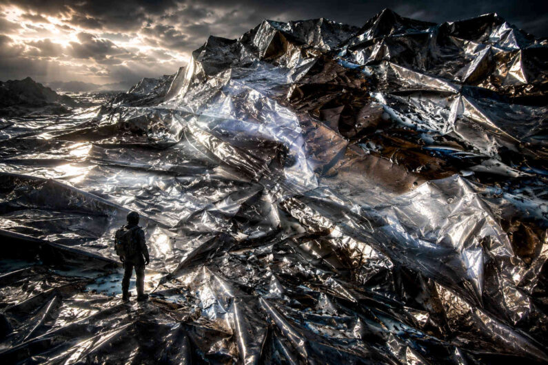 Gewaltige Berge aus glänzendem, metallisch reflektierendem Material dominieren die Landschaft. Eine einzelne Person betrachtet die zerknitterten Oberflächen unter dramatischem Himmel. Lichtstrahlen brechen sich in zahllosen Falten und Kanten. Das Bild erzeugt den Eindruck einer Welt aus flüssigem Metall.