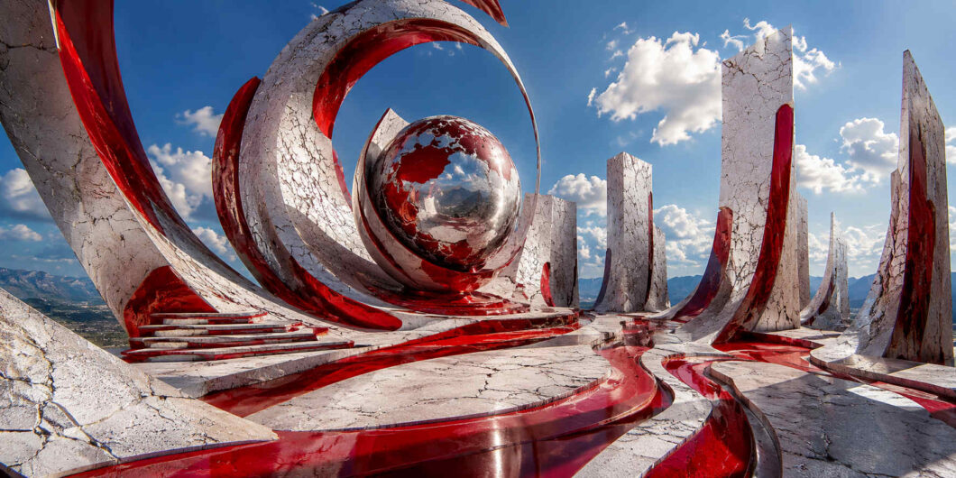 Monumentale rote und weiße Formen umgeben eine große spiegelnde Kugel im Zentrum der Komposition. Geschwungene Bahnen führen durch eine offene Anlage aus hohen Säulen und Bögen. Die glänzenden Oberflächen reflektieren Himmel, Wolken und Landschaft. Das Werk verbindet skulpturale Architektur mit symbolischer Wirkung.