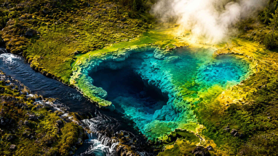 Luftaufnahme einer intensiv türkisfarbenen heißen Quelle mit tiefem dunklem Zentrum. Umgeben von moosbewachsenen Felsen, dampfenden geothermalen Feldern und einem Bachlauf. Die leuchtenden Mineralablagerungen erzeugen starke Farbkontraste zwischen Grün, Blau und Gelb. Die Szene wirkt wie ein verborgenes Naturwunder aus einer fremden Welt.
